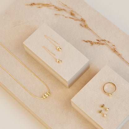 Collectie Essentials: 14 karaat gouden  minimalistische organische gouden steentjes aan een ketting, oorhangers, oorbellen en een ring. Liggend op zandkleurige stenen met een gedroogd takje als decoratie. Basic dus, passend bij de betekenis van deze sieraden: minimalisme geeft je meer ruimte en aandacht voor wat echt belangrijk is in het leven. Essentials, meer is er niet nodig.