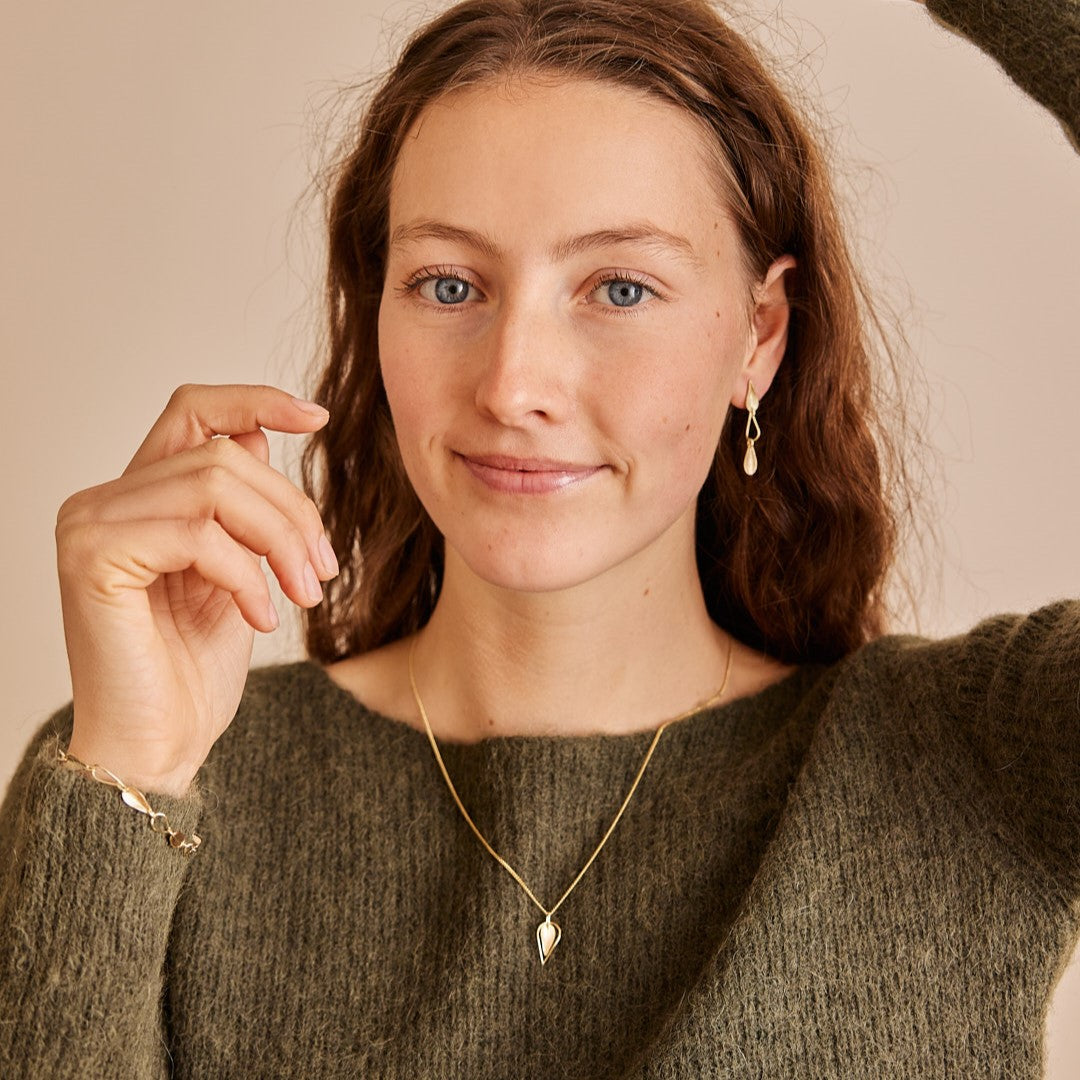 Model draagt het dubbele gouden hangertje Leaves om haar nek op een groene trui. Een minimalistisch gouden blaadje met daaromheen een open blaadje gemaakt van gouddraad als omlijsting.  Ook hoofd en armen van model zijn te zien waarmee ze de armband en oorhangers uit zelfde collectie laat zien.