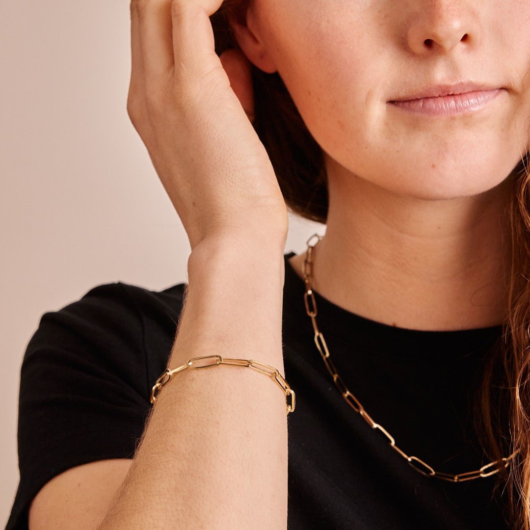 Model met zwart t-shirt draagt een gouden schakelarmband en schakelketting van de collectie Verbondenheid. De paperclipschakels staan symbool voor verbinding.